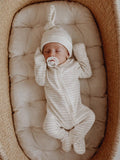 Baby in striped onesie and hat sleeps in a cozy basket, holding a pacifier on a cushioned surface.