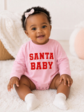 Smiling baby in a pink onesie with "SANTA BABY" text, sitting on a soft rug.
