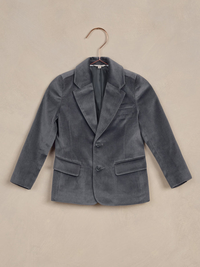 Gray velvet blazer hung on a copper hanger against a textured beige background.