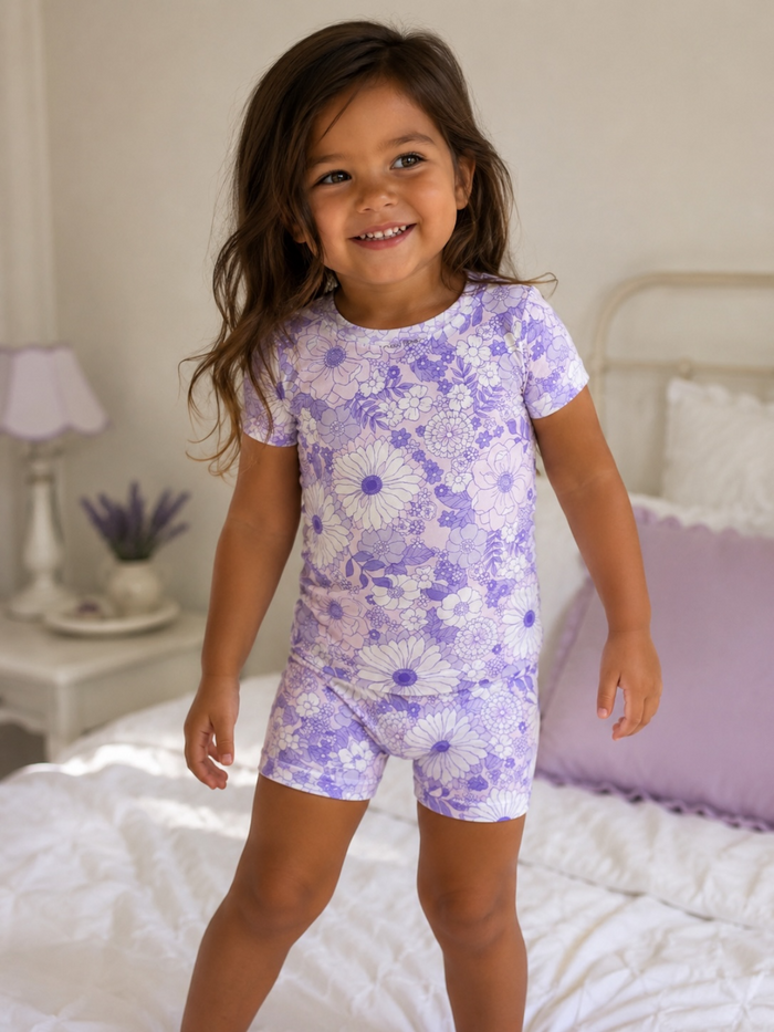Smiling child in floral purple pajamas, standing on a bed with soft decor in a bright bedroom.