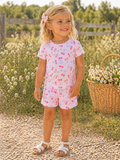 Smiling young girl in pink floral outfit, standing outdoors with greenery and a basket nearby.