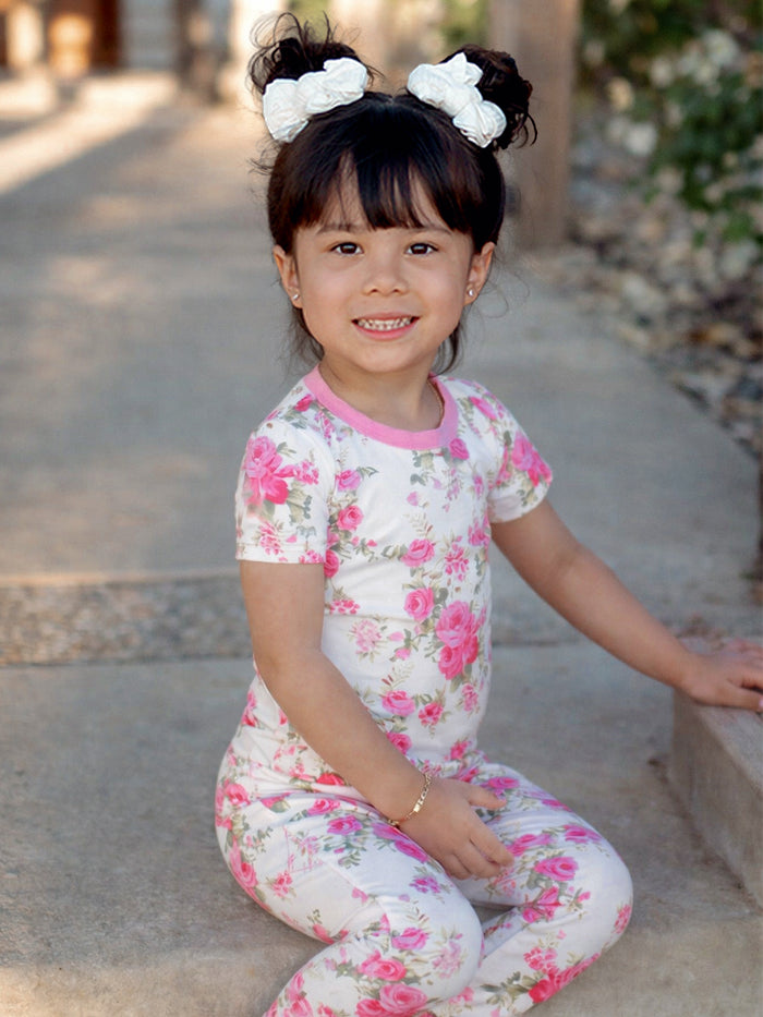 Smiling young girl in floral pajamas with two white bows in her hair, sitting on a pathway outdoors.
