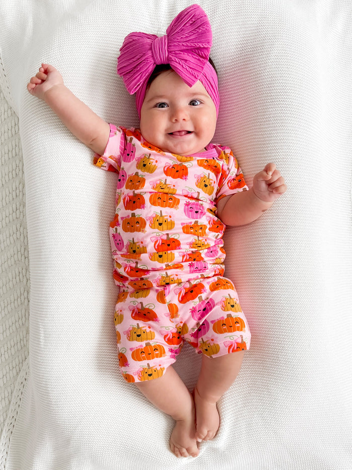 Smiling baby in a pumpkin-patterned outfit with a large pink bow, lying on a soft white blanket.