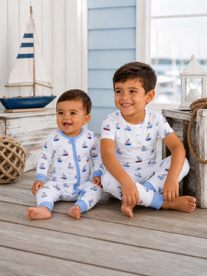 Two children in matching sailboat pajamas sit on a wooden floor, smiling, with nautical decor in the background.