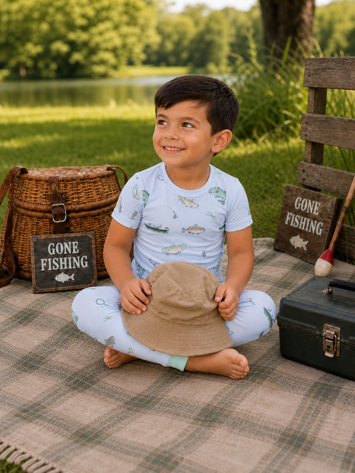 Smiling boy in light fishing-themed outfit sitting on a blanket by a lake, holding a hat with fishing gear nearby.