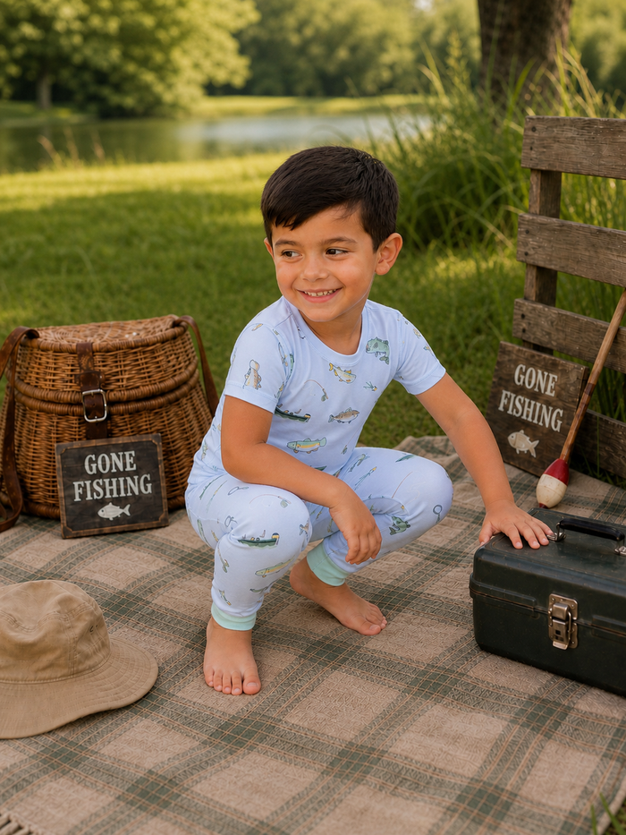 Smiling boy in fishing-themed pajamas crouches on a picnic blanket by a lake, surrounded by fishing gear and signs.