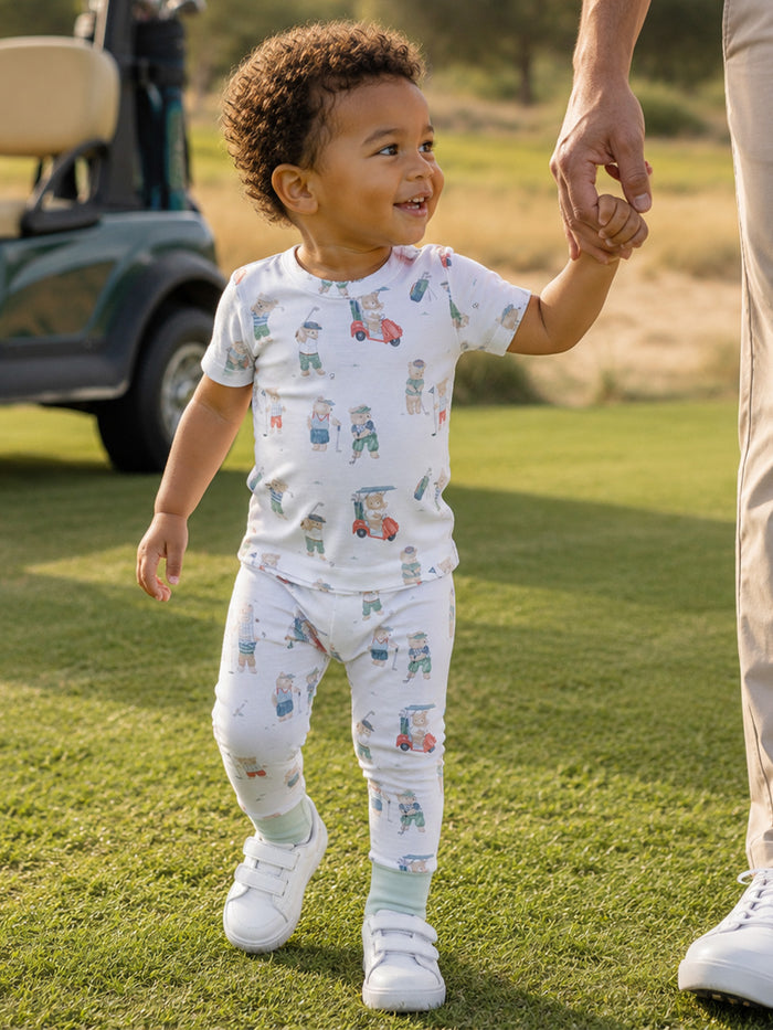 Smiling toddler in golf-themed pajamas walking hand-in-hand with an adult on a grassy field.