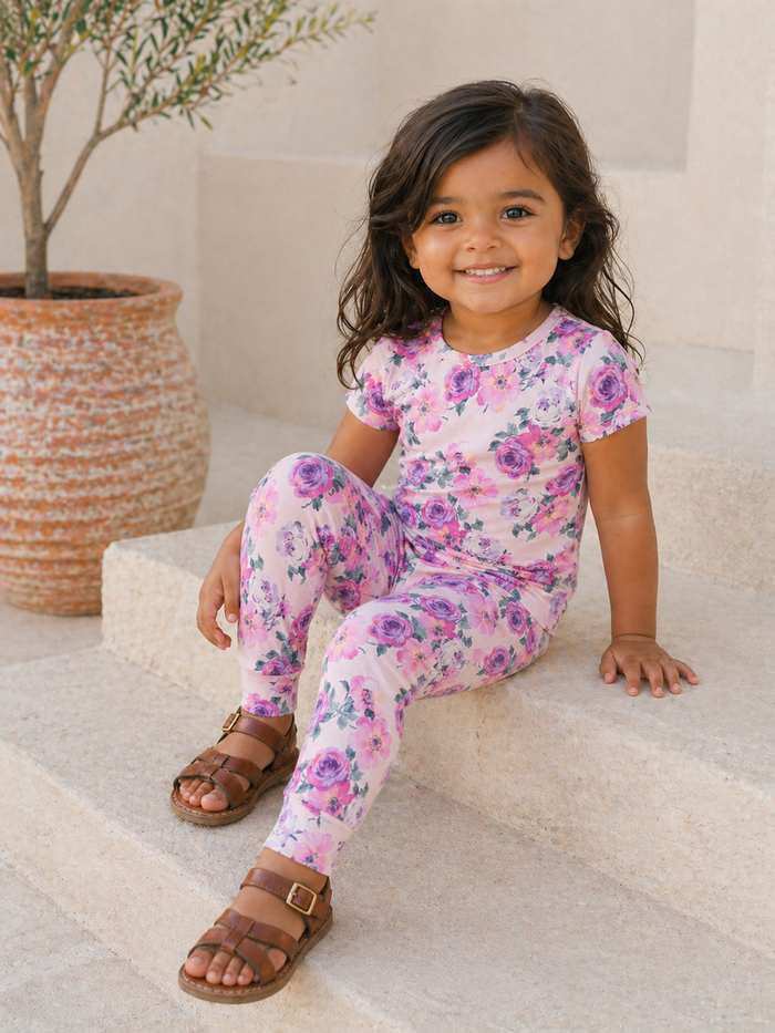 Smiling child in floral outfit sitting on steps near a potted plant, exuding joy in a sunny setting.