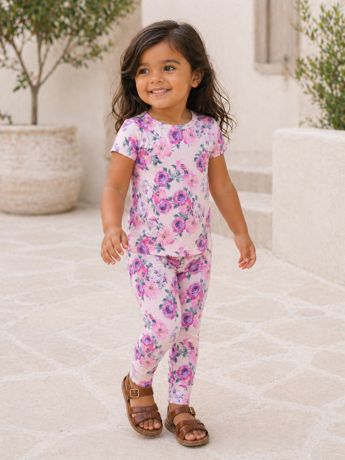 Smiling girl in floral outfit walks on stone path, with potted plants in background.