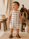 Smiling toddler in a plaid romper stands on a rug, with rustic decor in the background.