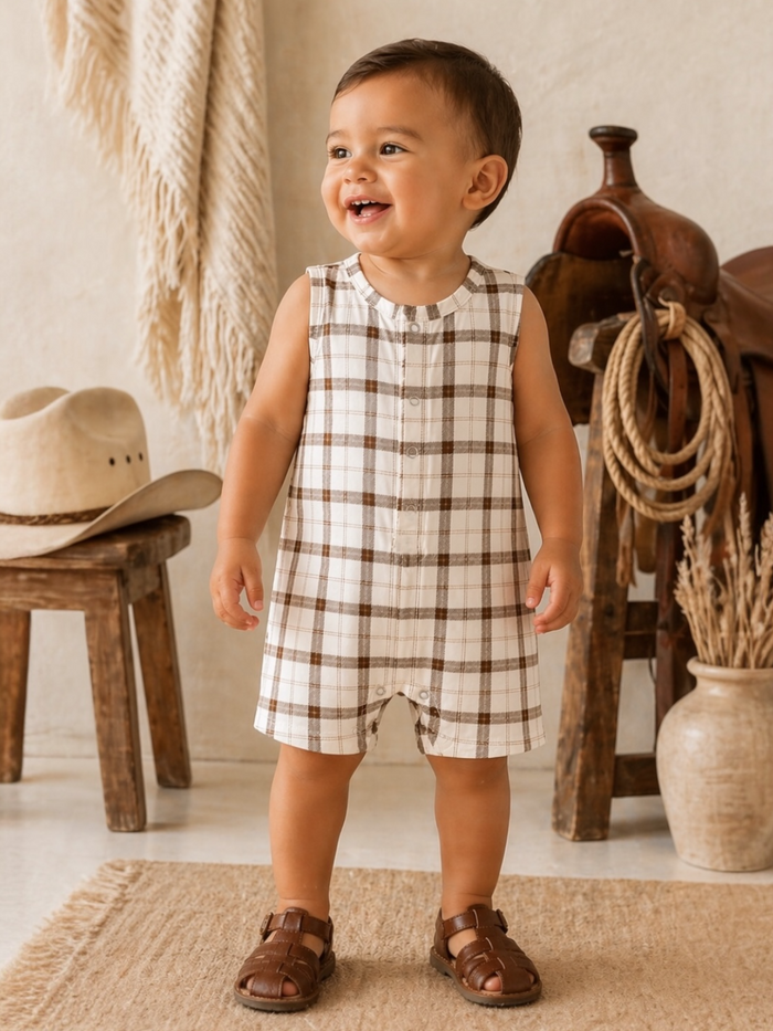 Smiling toddler in a plaid romper stands on a rug, with rustic decor in the background.