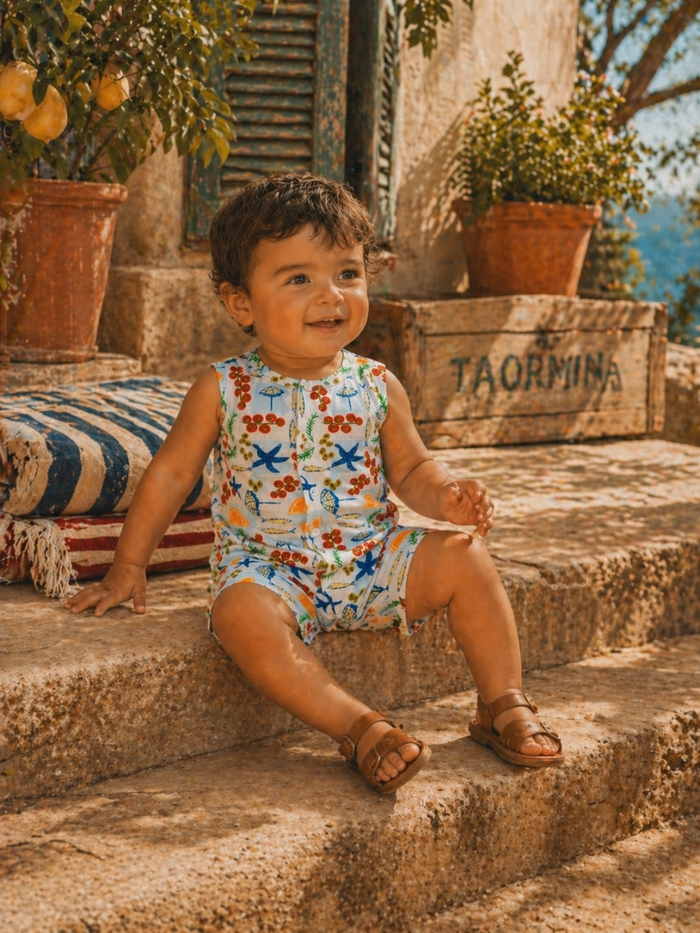 Smiling toddler in colorful outfit sitting on stone steps with greenery and rustic wooden box in background.