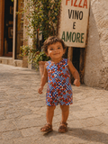 Smiling child in colorful romper stands on a cobblestone street near a pizza sign.