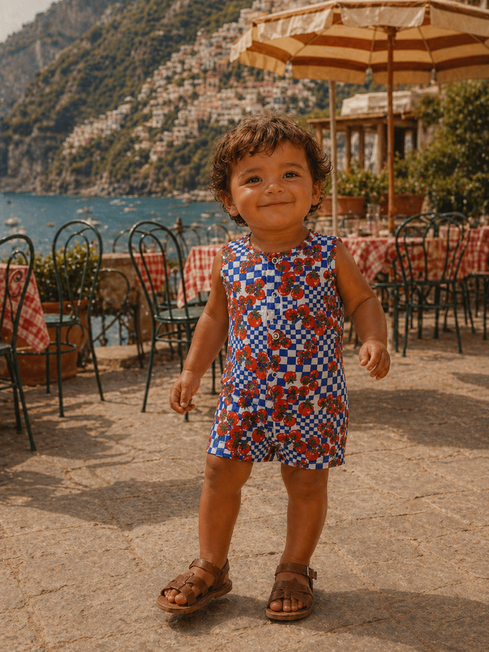 Smiling child in a colorful outfit stands outdoors near the coast, surrounded by tables in a picturesque setting.