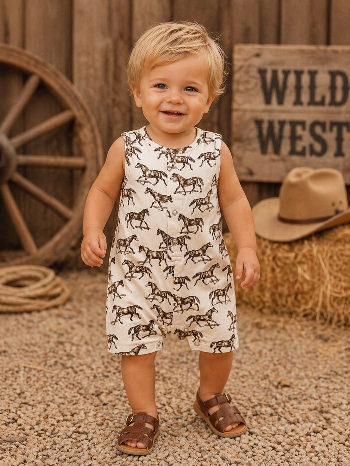 Smiling toddler in horse-patterned romper, standing on gravel with rustic backdrop.