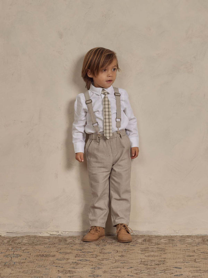 Child in beige pants, white shirt, and suspenders poses against a neutral background, showcasing a stylish outfit.