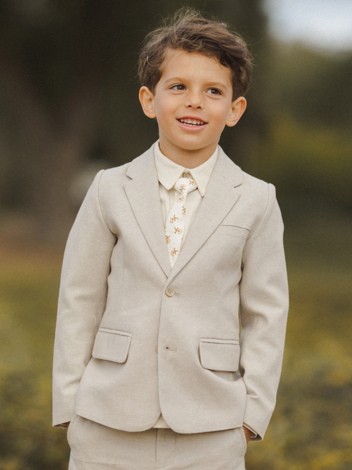 Smiling boy in a light suit with a patterned tie, standing outdoors in a natural setting.