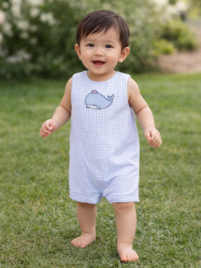 Smiling toddler in a blue gingham romper with a whale design, standing on lush green grass.