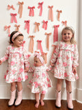 Three young girls in floral dresses holding hands, standing against a wall adorned with pink bows.