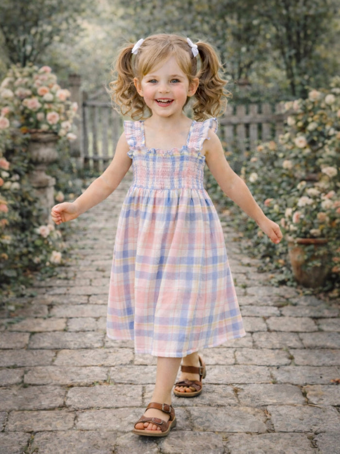 Smiling girl in a checkered dress walks on a stone path surrounded by flowers and greenery.