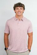 A smiling young man wearing a short-sleeve heathered pink polo shirt and khaki pants, standing against a plain light gray studio background.