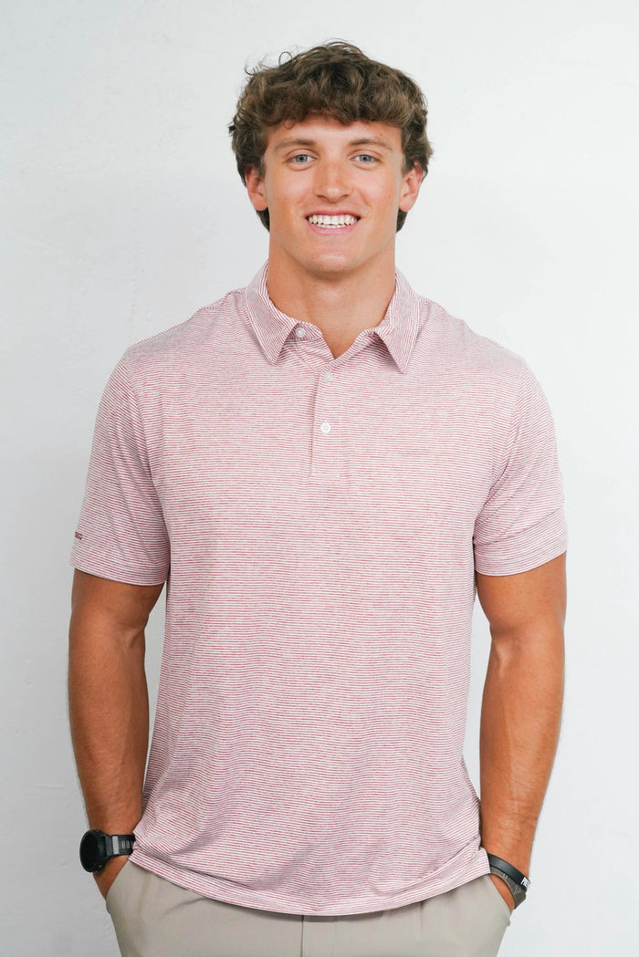 A smiling young man wearing a short-sleeve heathered pink polo shirt and khaki pants, standing against a plain light gray studio background.