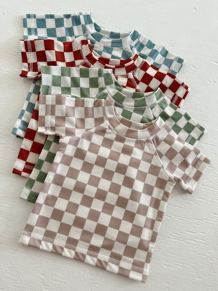 Four checker-patterned t-shirts in blue, red, green, and beige laid on a white surface.