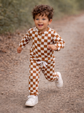 Smiling child in brown and white checkered outfit running on a dirt path surrounded by greenery.