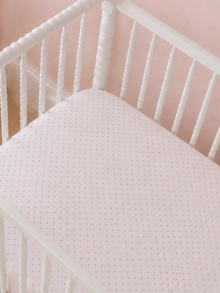 White crib with a pink polka dot mattress against a soft pink wall. Ideal for a nursery setting.