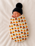 Newborn sleeping swaddled in pumpkin-patterned blanket with a black pom-pom hat on a soft white background.