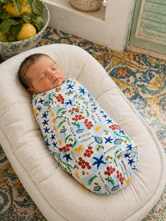 Sleeping baby swaddled in a colorful, patterned wrap on a soft surface. Background features lemons and patterned tiles.