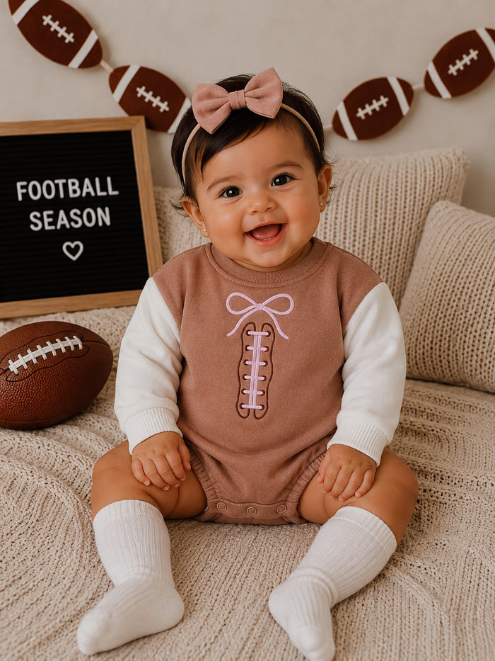 Smiling baby in a football-themed outfit with a pink bow, sitting on a cozy blanket with decor behind.