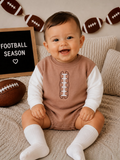Smiling baby in football-themed outfit, sitting on a cozy blanket near a football and "Football Season" sign.