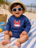 Smiling child in sunglasses, wearing a crab-themed outfit, sitting on a beach towel by the ocean.