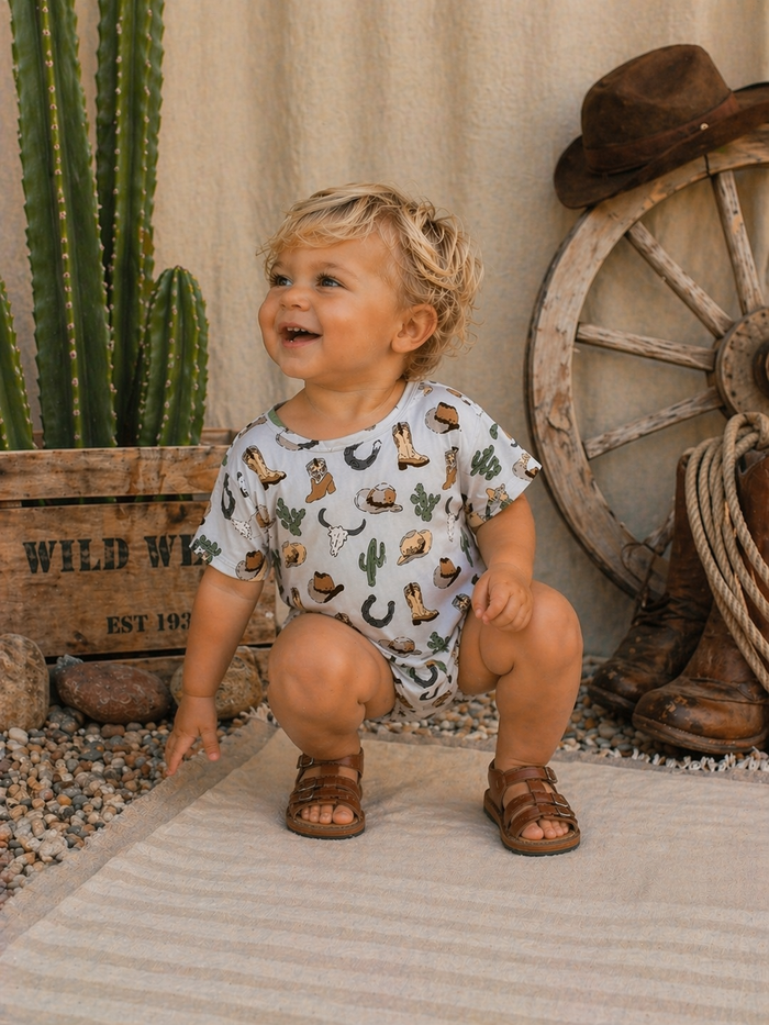 Smiling toddler in a patterned outfit squatting by cacti and rustic decor, capturing a joyful outdoor moment.