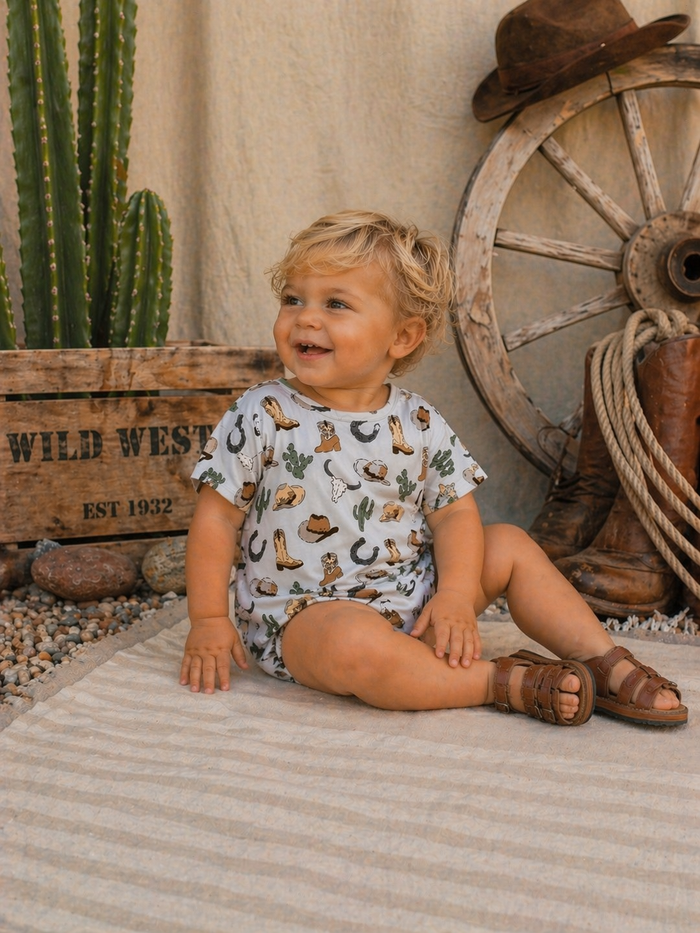 Toddler sitting on a rug, smiling, in a Wild West-themed outfit, with a cactus and wagon wheel in the background.