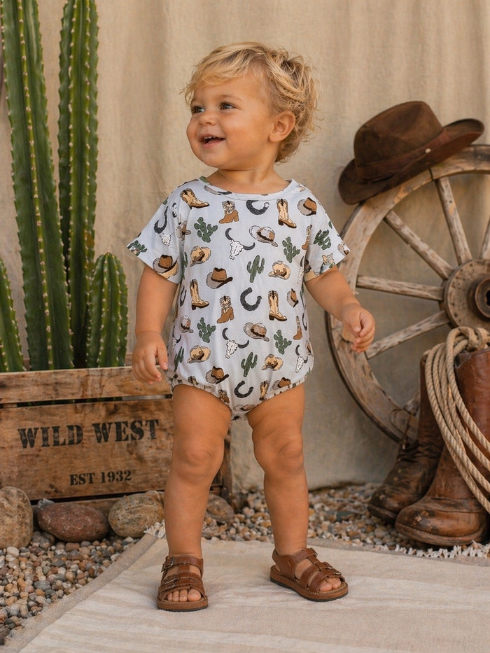 Toddler in a playful, western-themed onesie stands beside a cactus and rustic decor, smiling joyfully.