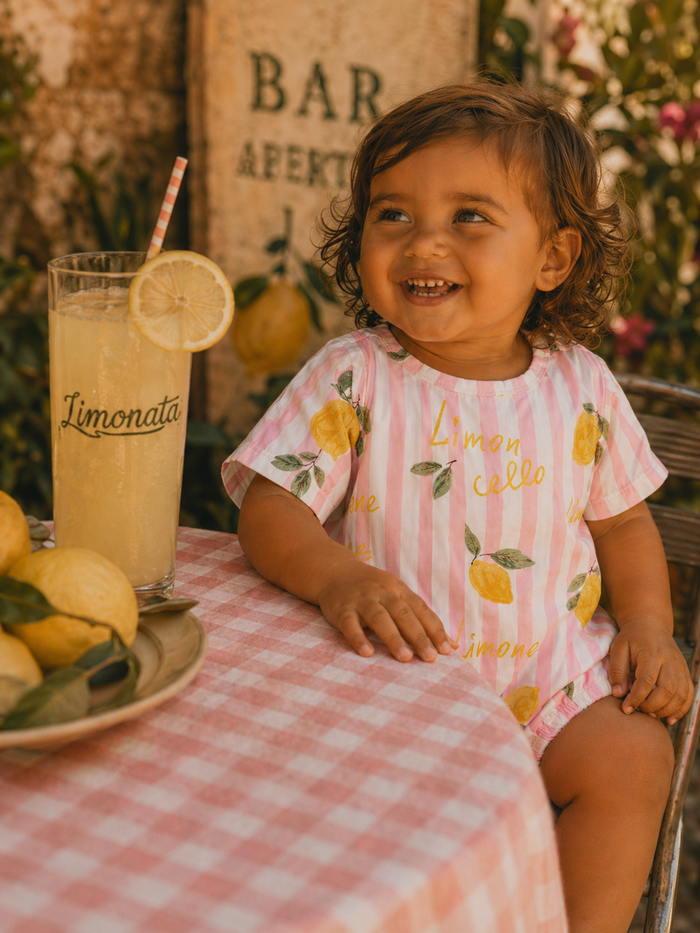 Smiling toddler enjoys Limonata at a table with lemons and a pink checkered tablecloth.