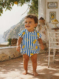 Smiling toddler in a lemon-patterned outfit stands outside in a sunlit, Mediterranean setting.