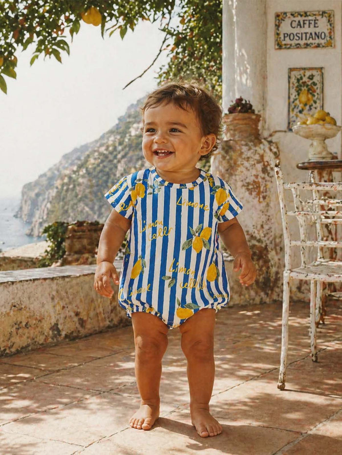Smiling toddler in a lemon-patterned outfit stands outside in a sunlit, Mediterranean setting.