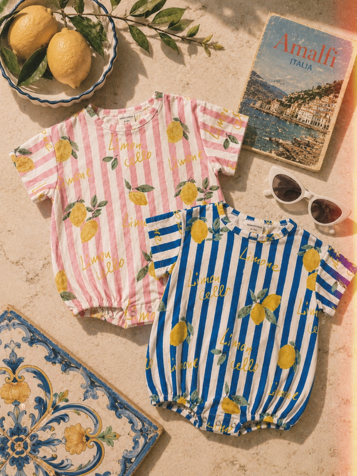 Two colorful baby onesies with lemon patterns on a stone surface, alongside lemons and sunglasses.