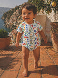 Smiling toddler in a colorful onesie, walking on a terracotta path with a scenic coastal backdrop.