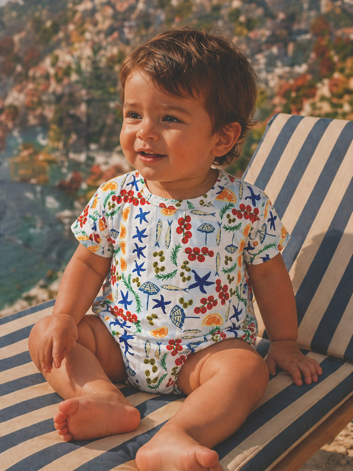 Smiling toddler in a colorful, patterned romper, seated on a striped lounge chair with a scenic background.