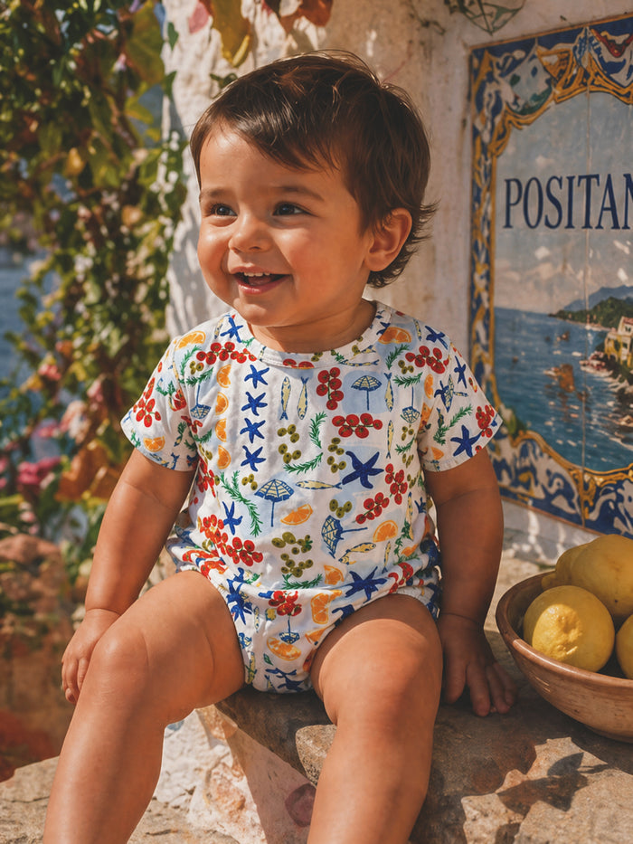 Smiling toddler in a colorful patterned outfit sits by a bowl of lemons and a mural in Positano.