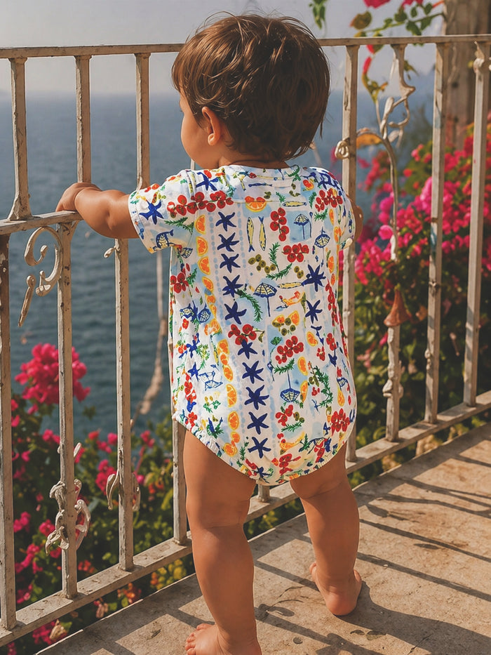 Child in colorful outfit gazes at the ocean, surrounded by flowers and a railing. A sunny day by the seaside.