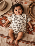 Smiling baby in a horse-printed onesie, lying on a patterned blanket beside a hat and rope.