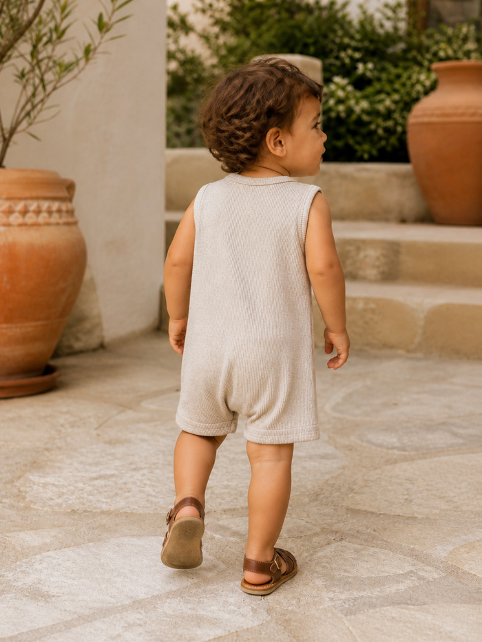 Toddler in a light romper walking away on a stone pathway, with greenery and terracotta pots in the background.