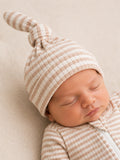 Sleeping newborn wearing a striped hat and sleep suit, resting peacefully on a soft, neutral-colored surface.