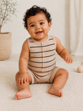 Smiling baby with curly hair, wearing a striped outfit, sitting on a soft rug with decor in the background.
