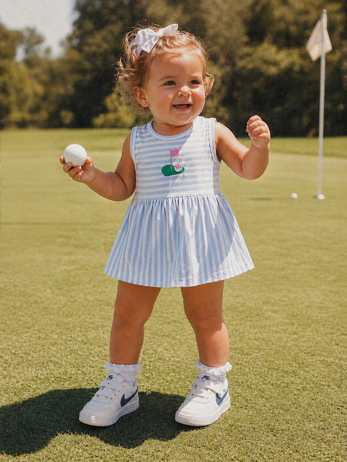 Smiling toddler in a striped dress holds a golf ball, standing on a green field with a golf flag in the background.